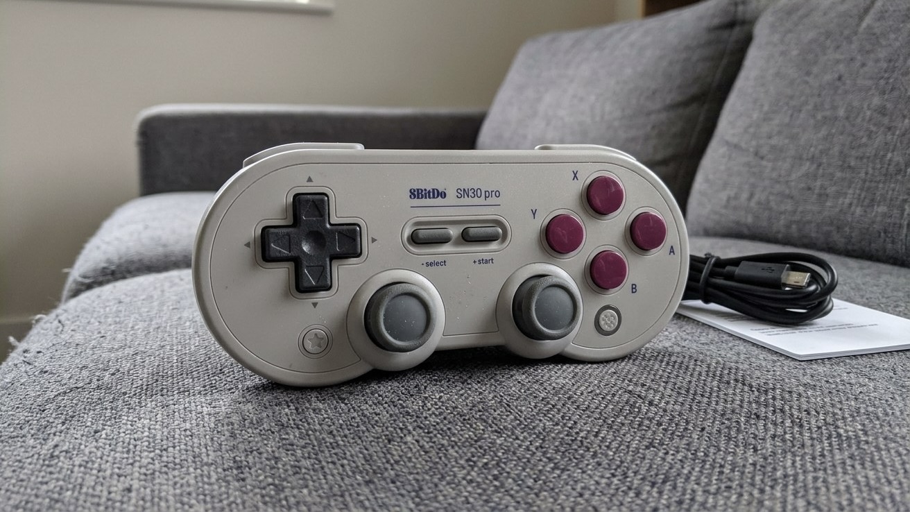 Серый геймпад 8BitDo SN30 Pro с фиолетовыми кнопками на диване, рядом кабель USB-C