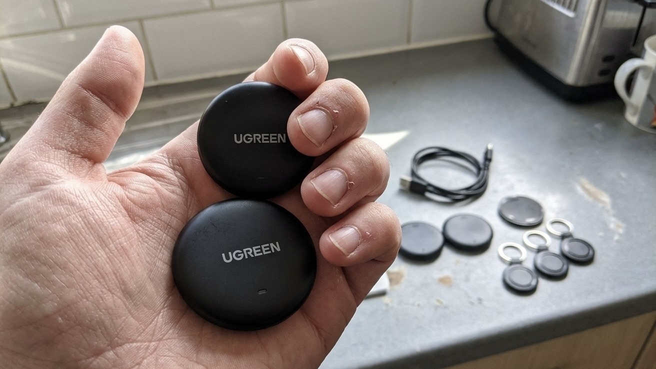 Два круглых трекера UGREEN в руке, на столе еще несколько меток, брелоки и кабель для зарядки