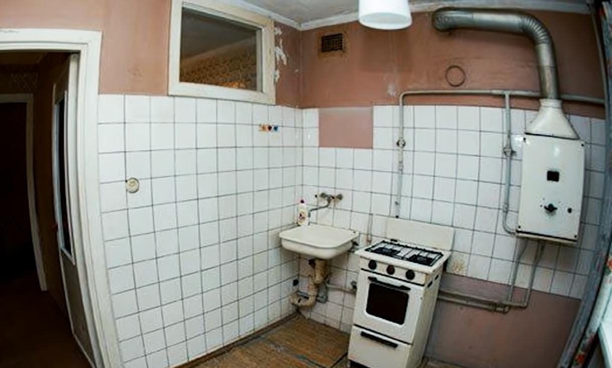 Старая советская кухня с газовой колонкой, открытыми трубами и небольшим окном в соседнее помещение.